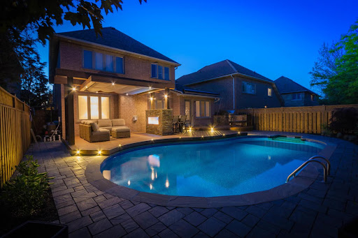 A landscaped backyard with an inground pool, patio, and covered deck with lighting.