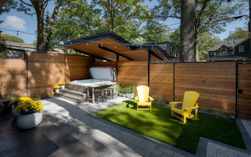 A backyard with a covered hot tub, wood fence, stone patio, and turf area.