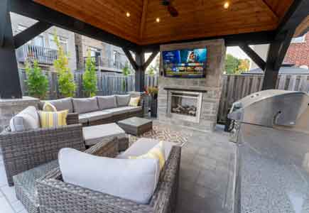 Covered pavilion backyard with outdoor kitchen, stone fireplace wall, putting green, and lighting in a narrow access Toronto townhome