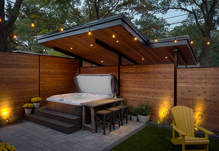 Modern Toronto backyard with dual-purpose roof covering hot tub and carport, stone patio, artificial turf, and warm evening garden lighting.