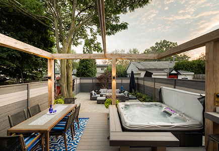 Cozy downtown Toronto backyard featuring a PVC deck, timber pergola with ShadeFX awning, hot tub seating, stone patio with fire table, and surrounding garden beds.
