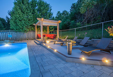 Caledon backyard with two decks, poolside stone patio, pergola with retractable awning, gazebo seating, cedar shed, and warm evening lighting.