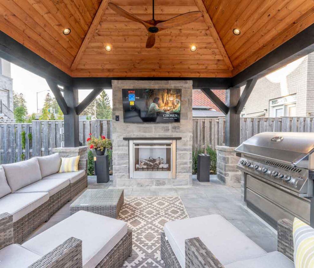 Cabana with a vaulted ceiling featuring a statement wall with TV and fireplace, paired with a comfortable lounging area and an integrated outdoor cooking station.