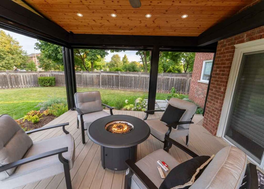 Deck with a roof cover and fireplace, surrounded by landscaped gardens and lush green space, featuring retractable screens for shade during the day and bug protection at night.