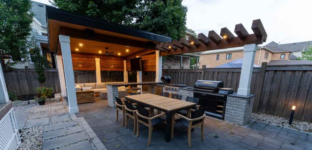 “Fully landscaped backyard featuring a dining area, custom outdoor kitchen, pergola with integrated lighting, and a cabana creating a complete outdoor living space for summer entertaining.