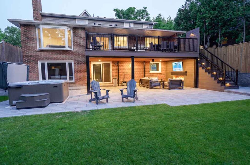 Fully landscaped backyard featuring a second-floor deck with a basement walkout and a covered seating area below, complete with a feature wall, fireplace, and evening lighting