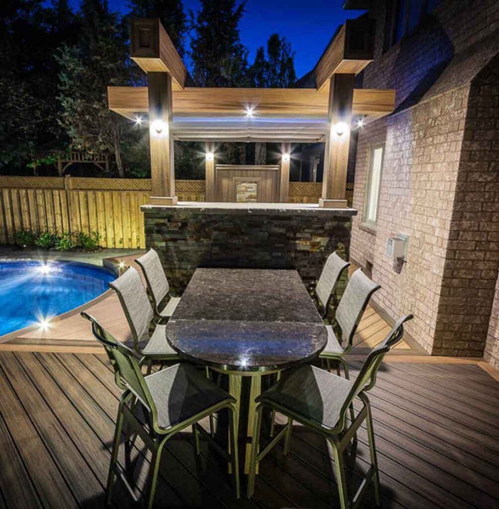 Night view of a poolside deck with a well-lit outdoor dining area and a pergola in the background
