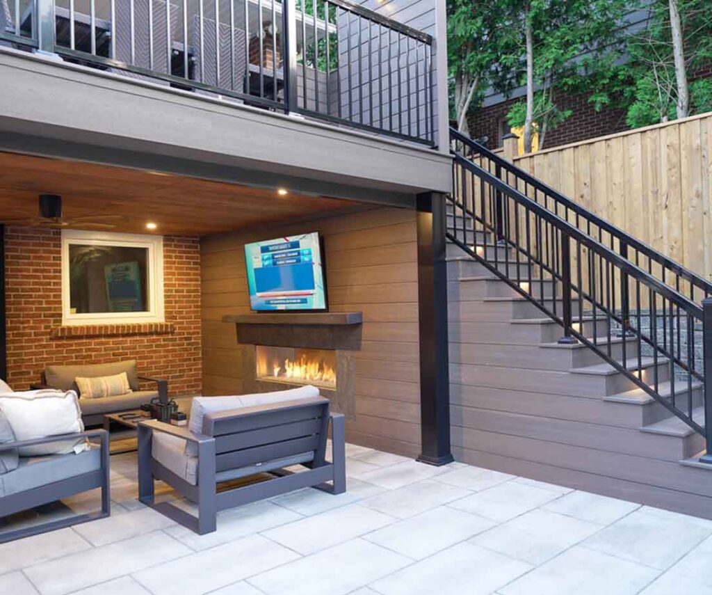 Second-floor deck designed as a roof, creating a sheltered living space below with a lounge area, feature wall with fireplace and TV, and integrated storage, all combined into one seamless outdoor living space.