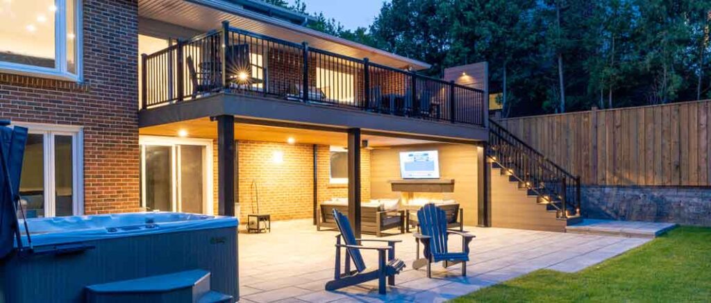 “Backyard landscaping with a hot tub and seating area beneath a waterproofed second-floor composite deck featuring a decorative feature wall.