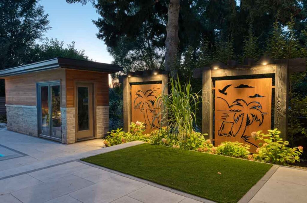 Custom modern shed with stone and wood finishes, surrounded by porcelain tile, privacy screens, and lush tropical-style planting.