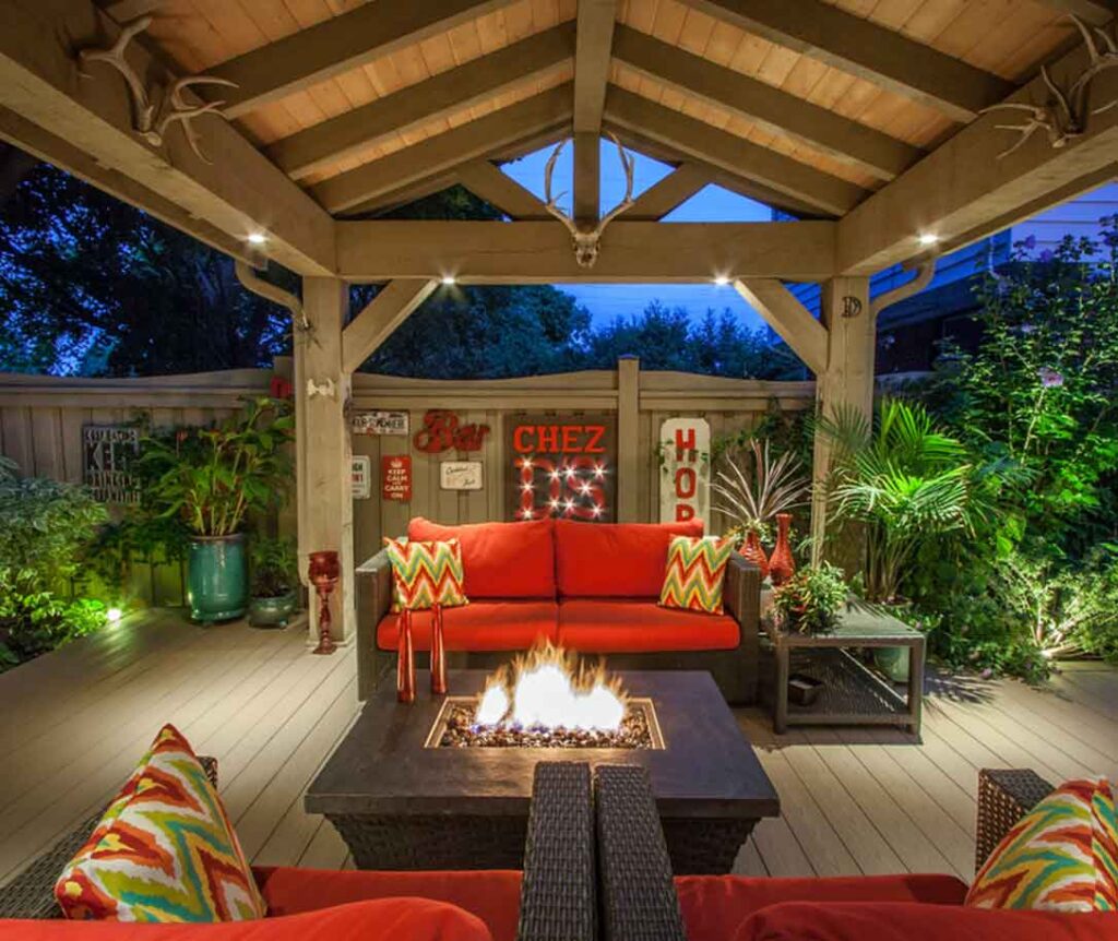 Timber cabana at dusk with a fire table beneath the roof, surrounded by lush greenery and designed as a relaxed staycation-style outdoor retreat.