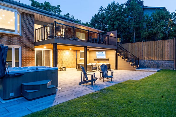 Backyard with large stone patio and hot tub, plus waterproof second-floor deck creating a dry lounge area below with fireplace feature wall, TV, and storage under stairs.