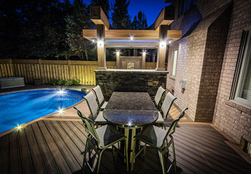 Curved Trex deck surrounding a kidney-shaped pool with fire pit, pergola with fireplace, outdoor kitchen, and bar seating