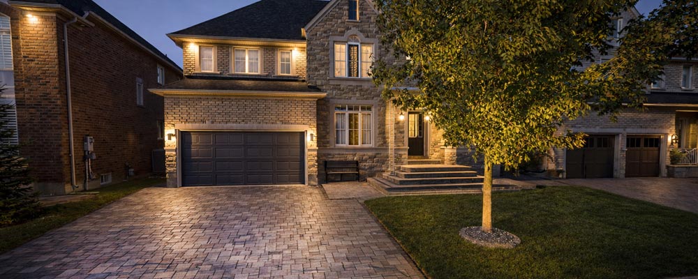 Front yard interlock driveway installed over concrete base for durability with stone porch and landscape lighting at night.