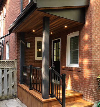 Modern covered front porch in Toronto with composite decking, black aluminum railings, brick facade and cedar soffit ceiling with recessed lighting