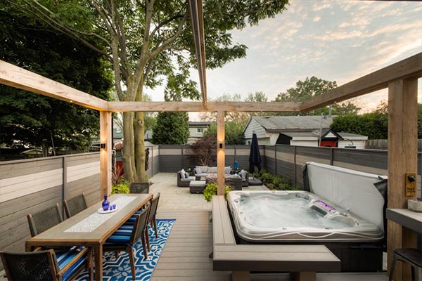 Modern Toronto Beaches backyard with large timber pergola and retractable awning, PVC deck with hot tub, and lower stone patio lounge with fire table under a mature tree.