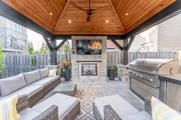 Backyard pavilion with stone outdoor fireplace, TV above, lounge seating and outdoor kitchen in a townhome backyard